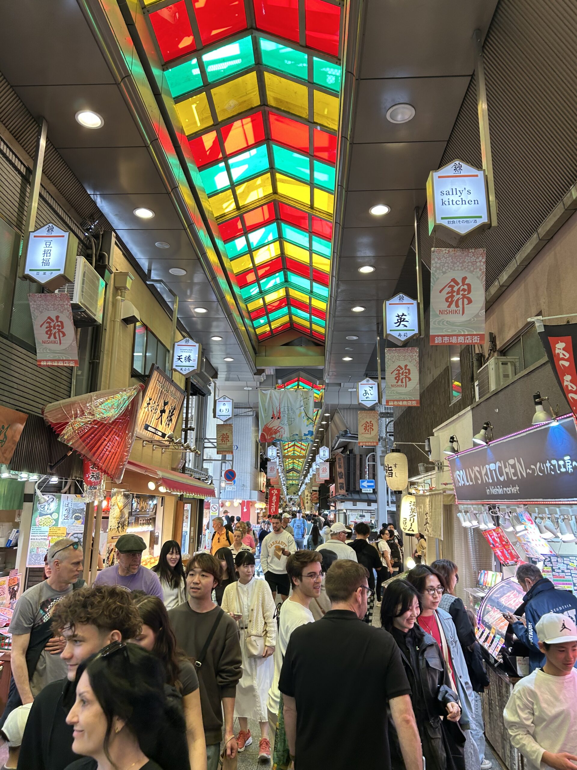 The covered arcade of Nishiki Market in Kyoto, with a coloured glass ceiling above the crowd.