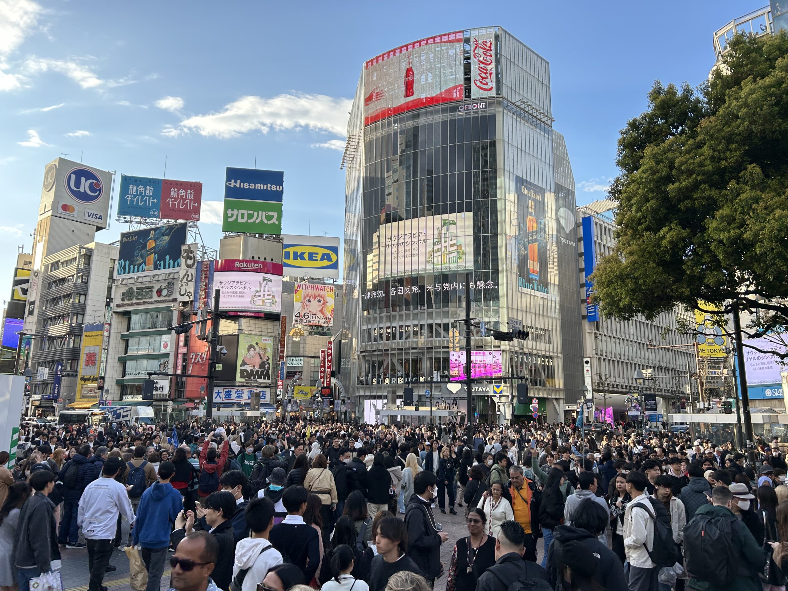 Crowds filling Shibuya Crossing in Tokyo beneath advertising screens and a blue sky.