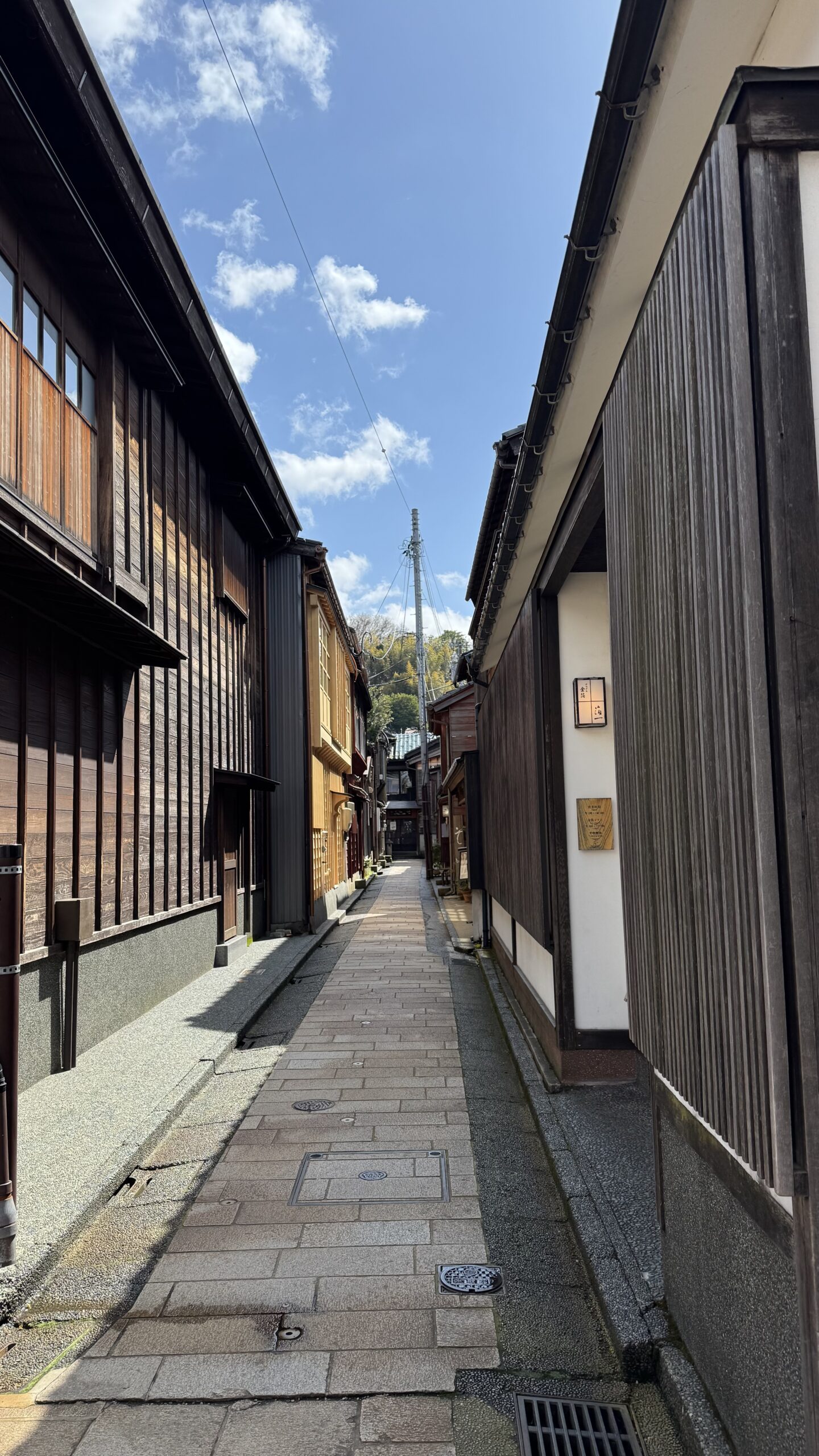 An empty lane of wooden merchant houses in the Higashi Chaya district of Kanazawa under a blue sky.
